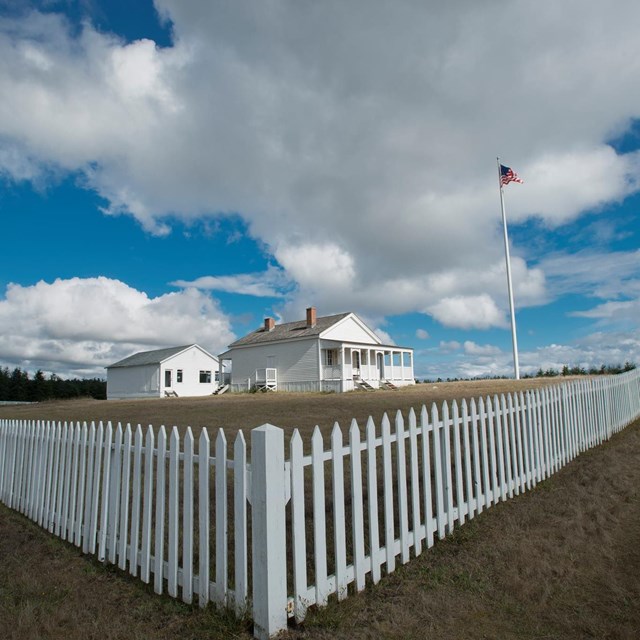 A white picket fence surrounds a grassy field with white historic buildings