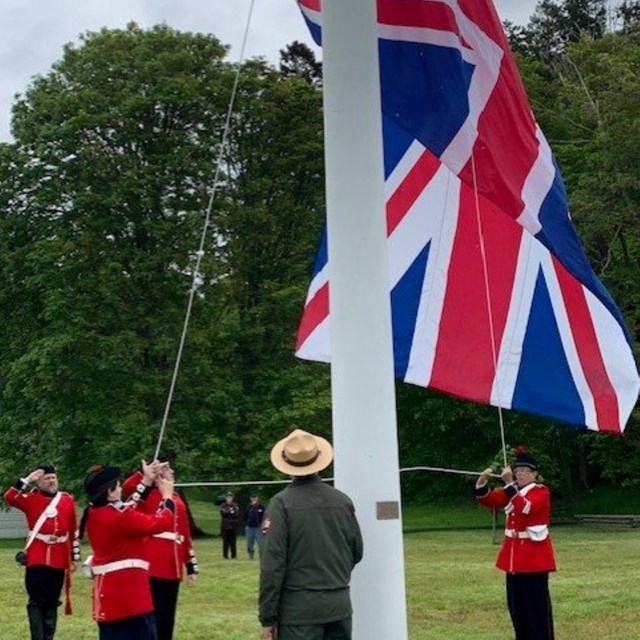 Color photo of a park ranger watching men in red coat uniforms raise an English flag