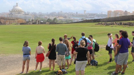 Park Ranger leads group in outer defenses of fort. Puerto Rican capitol building is seen behind.