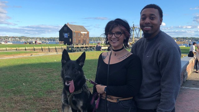 A man and woman stand with their dog in front of Salem Maritime's wharf.