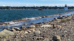 rocky shoreline with high winds making waves in the harbor and lighthouse and ships in the distance