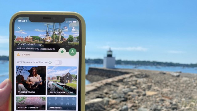 A phone showing Salem Maritime on the NPS App is held up with a lighthouse in the distance.