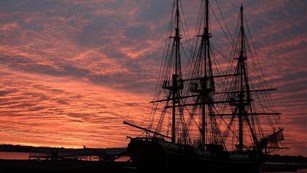 The sun sets on a large three-masted tall ship. 