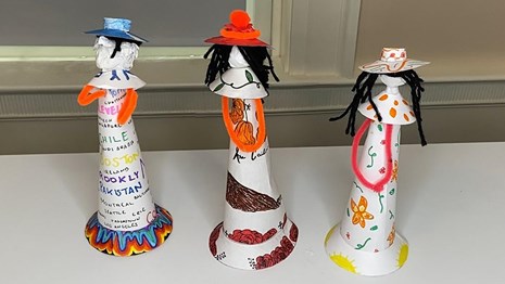 Three paper faceless dolls with black hair, colorful hats, and patterned dresses.