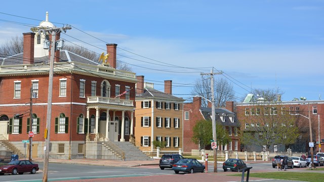 Cars travel down the road in front of multi-story historic buildings.