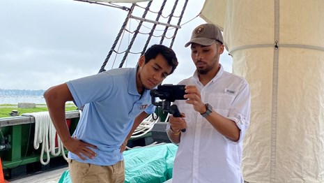 Two interns stand on a ship looking at a camera recording on a phone.