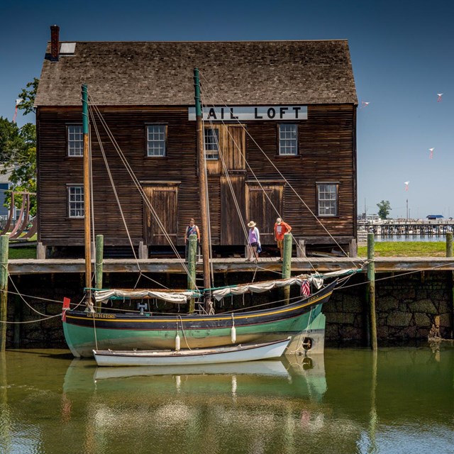 A sailboat is in front of a two story wooden building on the edge of the water.