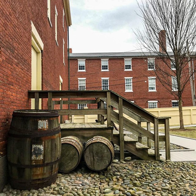 barrels are in front of wooden stairs which lead up to the closed door of a brick building.