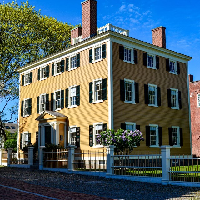 yellow three-story federal style home with rows of windows with green shutters and a fence