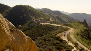 A dusty trail extends into the distance along a green mountain ridge
