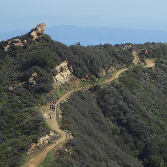 Hikers in the distance hiking along a mountain trail with the pacific ocean in the background.