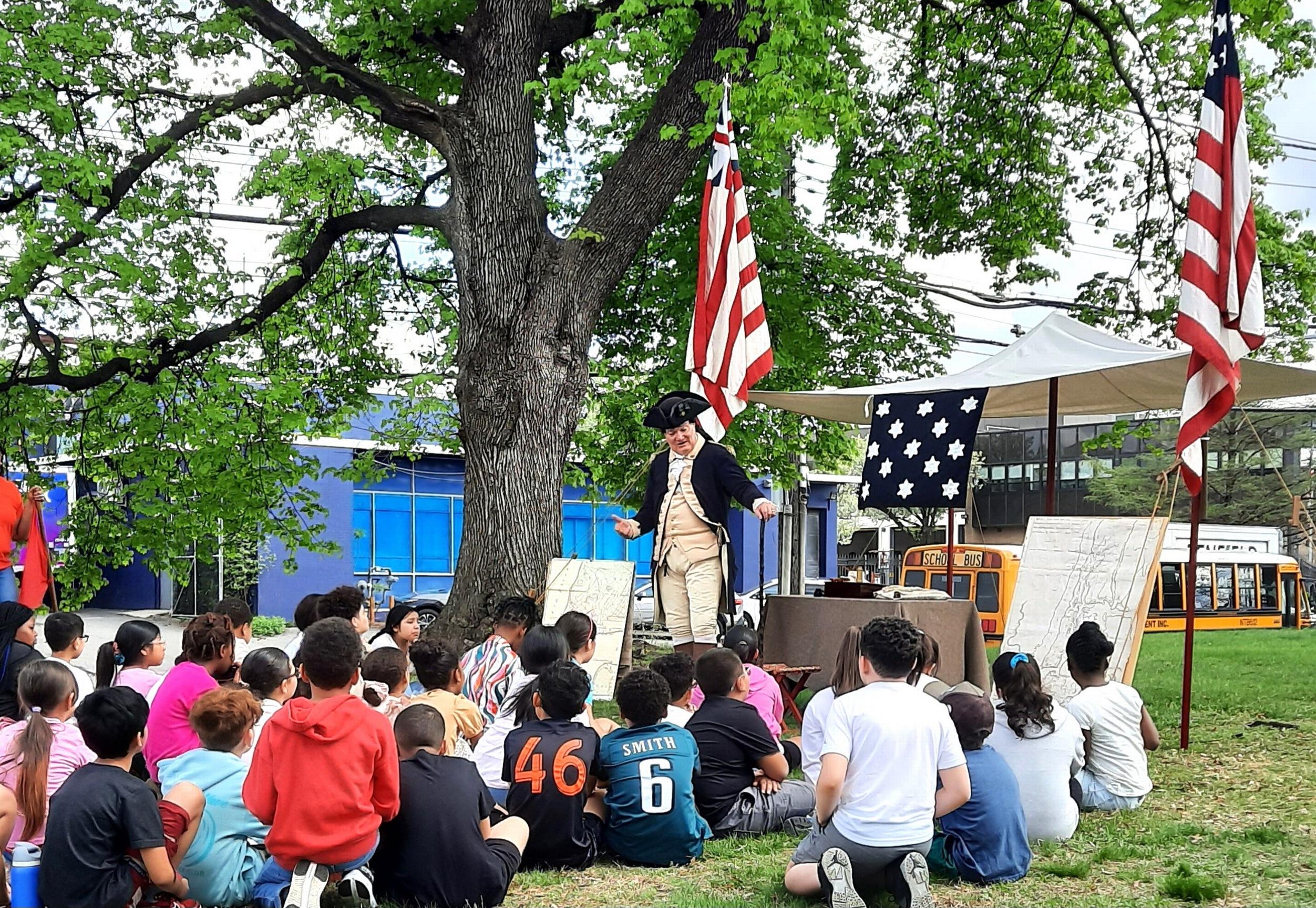 Students seated on a lawn looking at a Revolutionary War re-enactor, with flags, tree & tent.