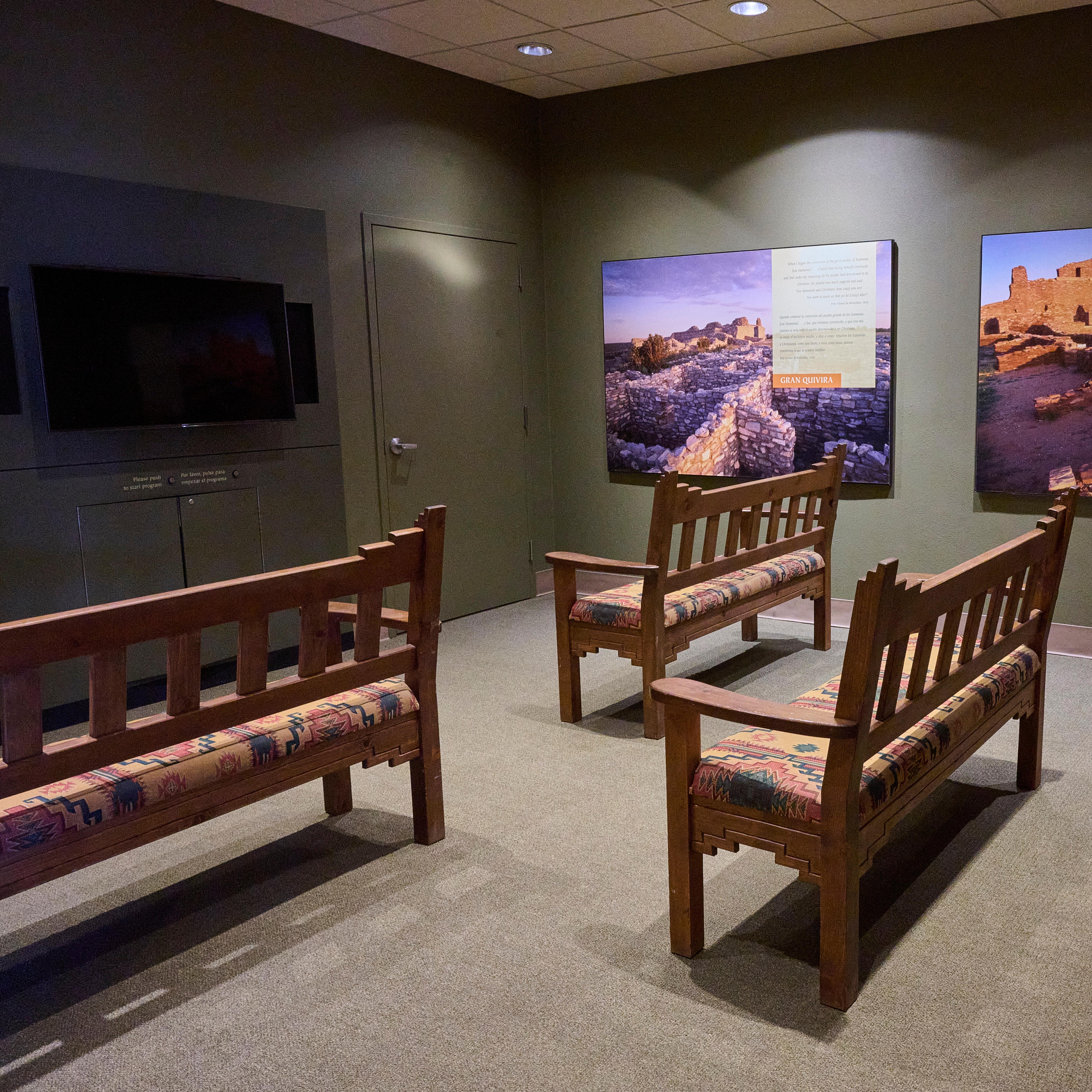 Small movie theater room with five wooden benches, television screen, and information panels.