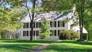 A pale yellow, two-story historic house surrounded by large trees and lush lawns.