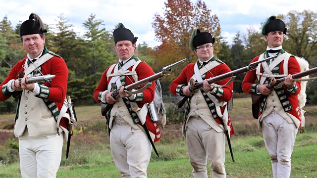 Four people dress in 18th century red uniforms with white pants and hold muskets.