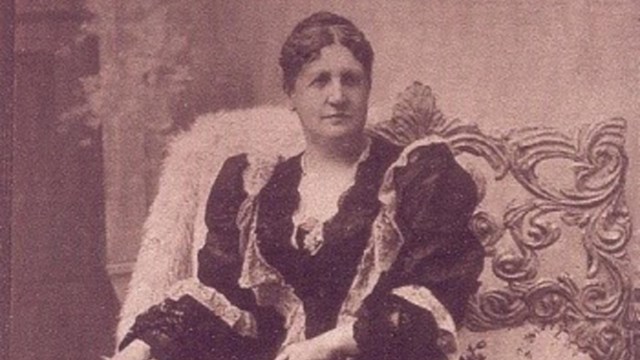 Middle aged woman in an ornate dress and seated on a chair with an open book on her lap.