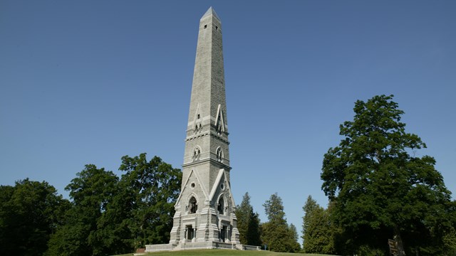 A clear blue sky over a tall gray obelisk monument with trees around the base.
