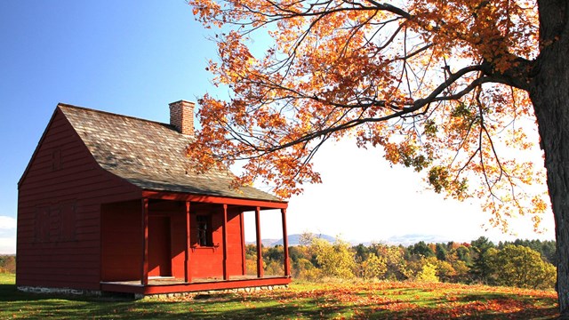 A red house sits on a hilltop surrounded by expansive views of hills, mountains, and fall foliage.