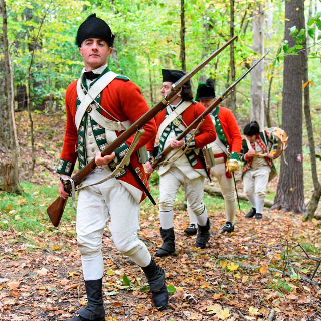 four soldiers wearing red coats marching on a trail in the woods while holding a musket