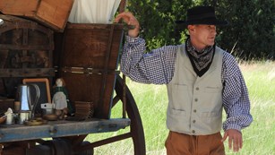 A man dressed in period clothing leans against a covered wagon.