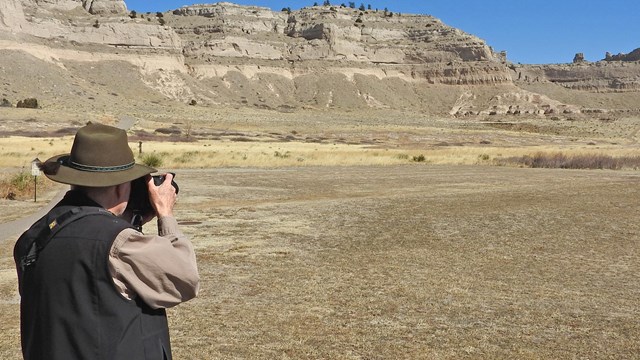A man holds a camera to his face overlooking a sandstone bluff. 