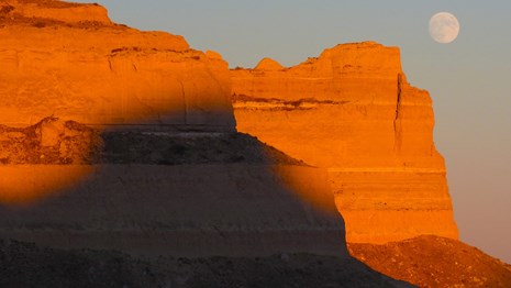Sandstone bluffs glow in orange light while the full moon is seen overhead. 
