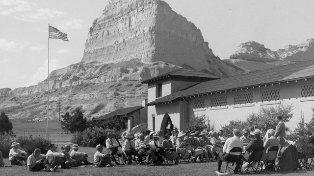 A black and white photo of people gathered below a sandstone bluff. 