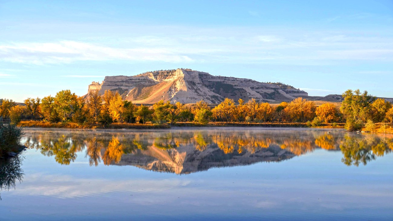 A sandstone bluff and trees with golden leaves are reflected in the waters of a pond.