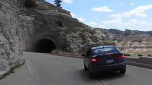 A gray car heads towards a tunnel carved in sandstone.