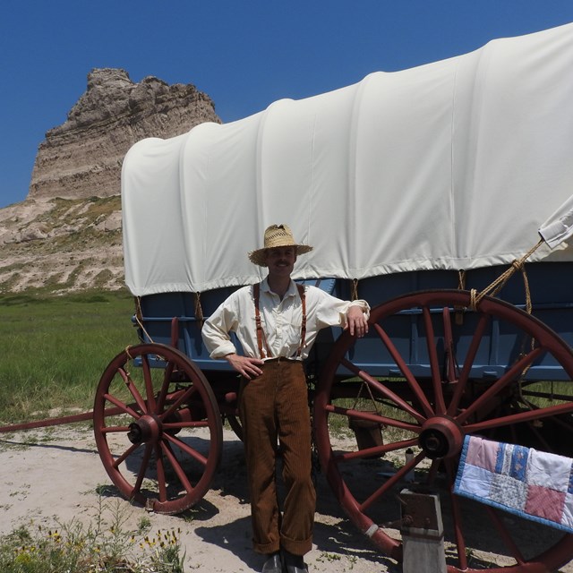 A man in period costume leans against a covered wagon.