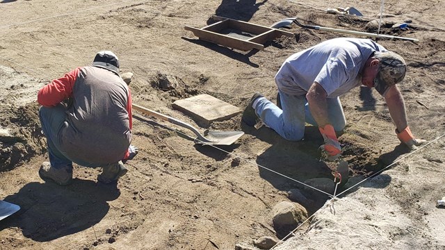 People leaning over in shallow pits, shovels and screens on the ground around them