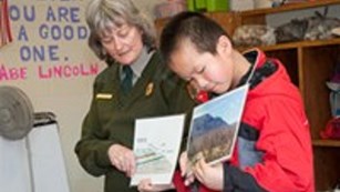 A woman with white hair in ranger uniform reads a book with a young boy in a classroom.