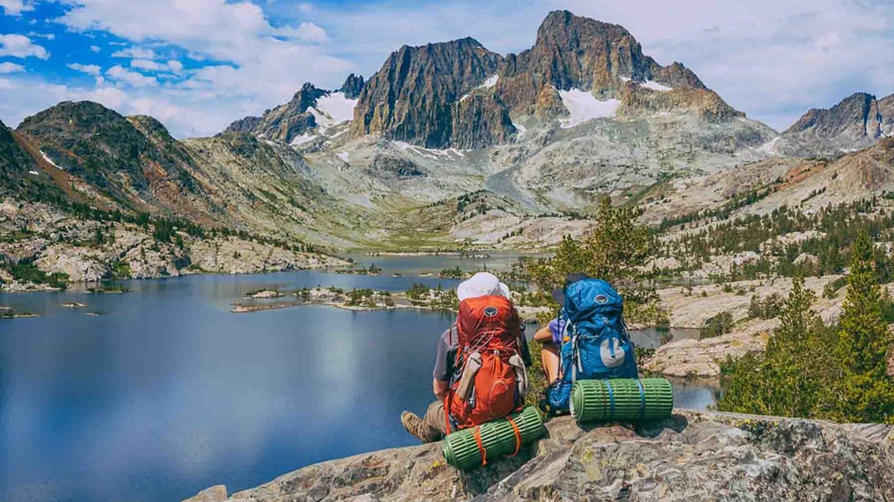 Two people sit on a high rock with their backpacks on looking over an alpine lake.