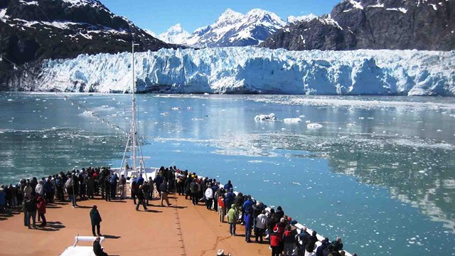 The bow of a cruise ship with people all along the railing looking at a tidewater glacier.