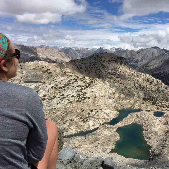 A woman sits with her back to the camera looking at an alpine lake.