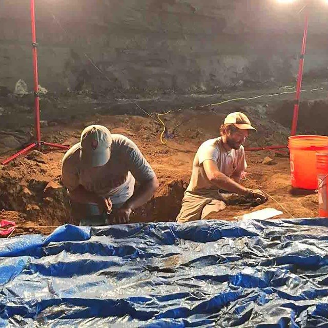 Two young people work on an excavation under lights.
