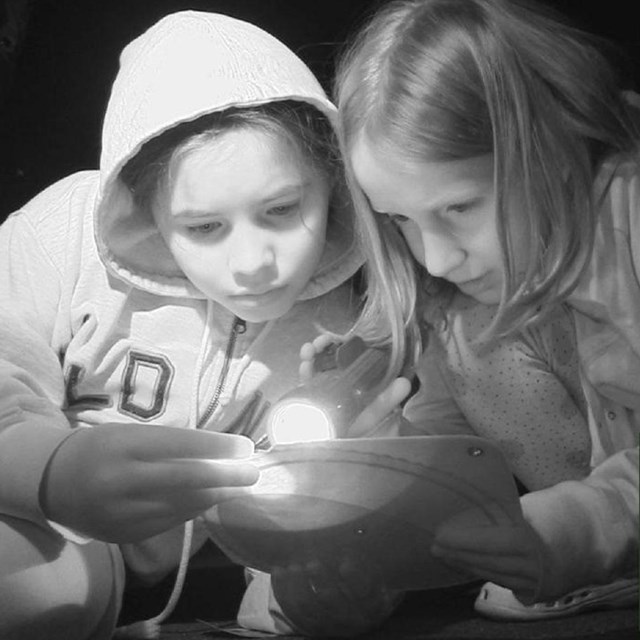 A black and white image of two girls huddled, reading a page with a flashlight