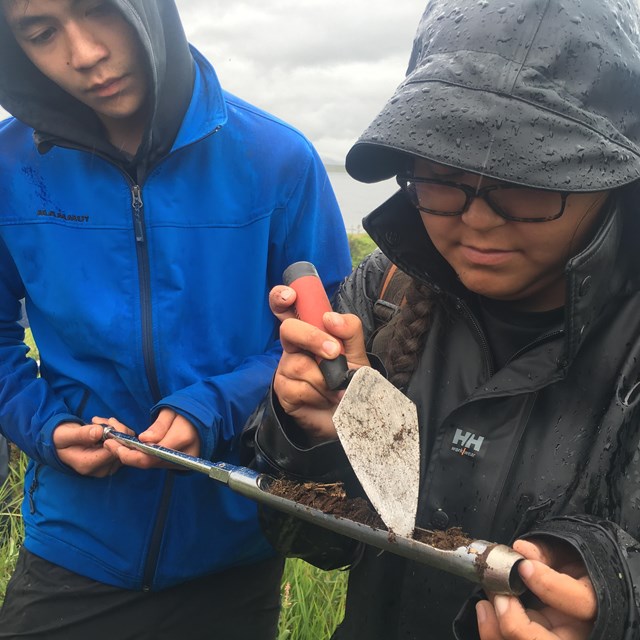 Two youth look at a metal rod with dirt in it; one poking it with a trowel
