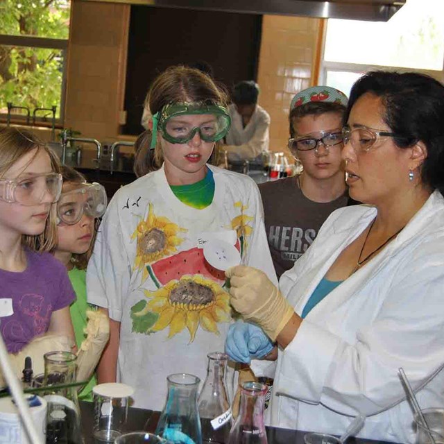 Three young girls listen to a woman scientist as she shows them something.