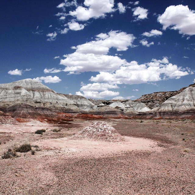 Barren badlands with striated hills in the distance under a blue sky with fluffy clouds.