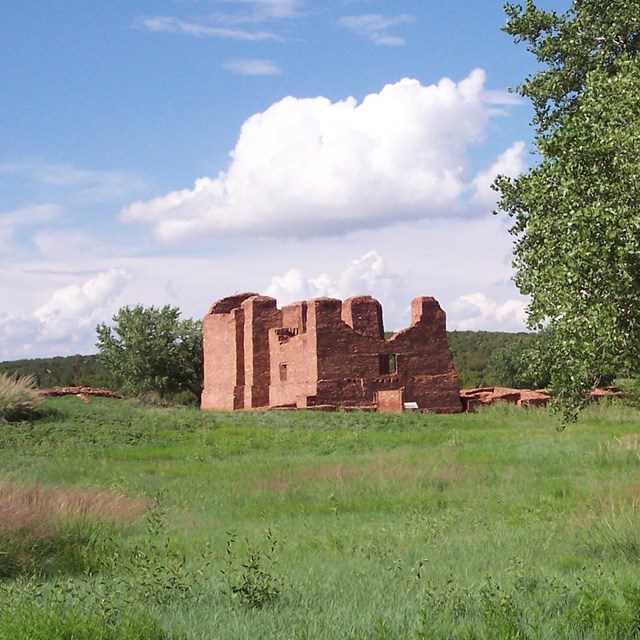 Ruins of a several story historic mission in a grassy meadow with a large tree on the right