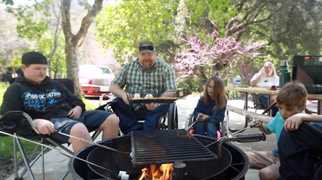 A group of adults and youth in wheelchairs and camp chairs roast marshmallows over a grill.
