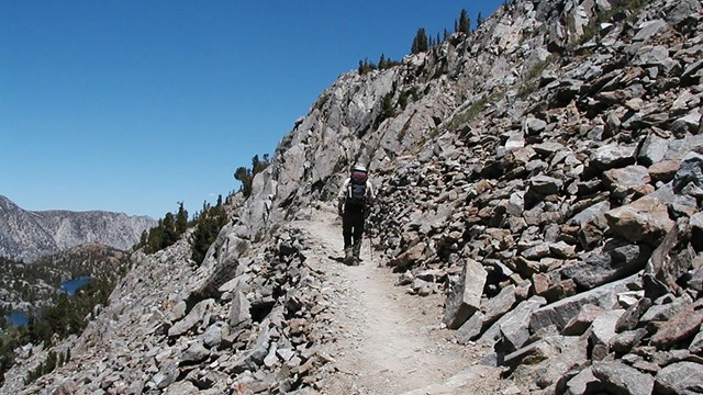 A backpacker walks into the distance along a trail cut into the side of a rocky slope.