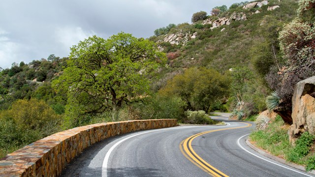 A two-lane road on a forested hillside with a rock wall guard rail winds into the distance 