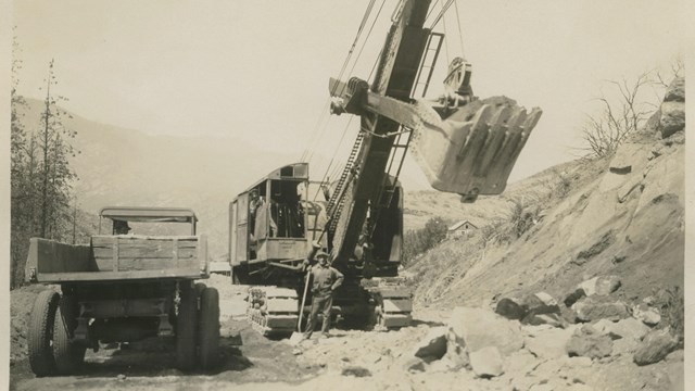 Historic black and white image of a steam shovel and a truck. The steam shovel digs into a hillside