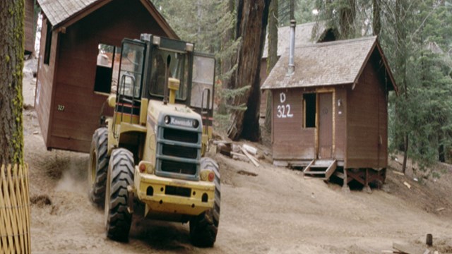 A yellow tractor moves toward a brown rustic structure.