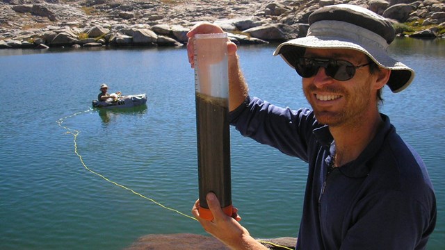 A researcher holds up a core sample near an alpine lake.