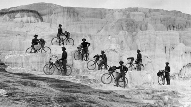 Eight dark silhouetted figures with bicycles posed on a hillside of light-colored travertine rock 
