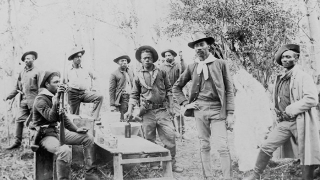 A black and white historic photo of Black male soldiers standing near a table in a forest