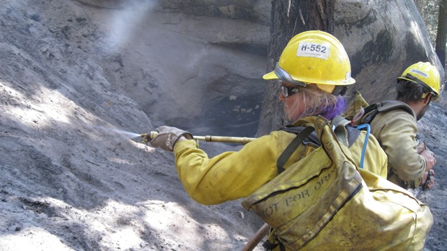 A woman in a yellow shirt and hardhat sprays water into a hot, ashy landscape.